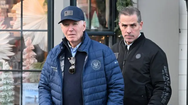 Joe Biden walking out of book store wearing blue jacket, jumper and shirt with cap emblazoned with US flag in blue. Son Hunter Biden is walking behind him in black jacket and white shirt