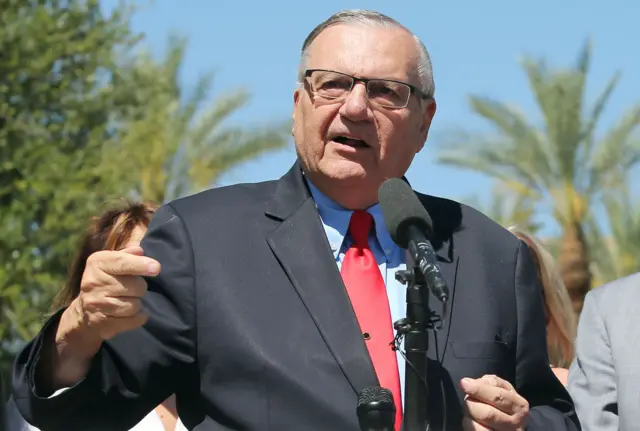 Joseph Arpaio in a suit with red tie, speaking into microphones with left hand raised and pointing away from him, with people in the background