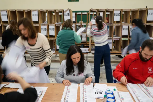 Count staff at Nemo Rangers GAA Club in Cork, as the vote count continues in the Irish General Election. There are four people around a table - three are sitting. They have ballot papers around them. There are four people behind them adding papers to stacked boxes which have excluded on a number of them