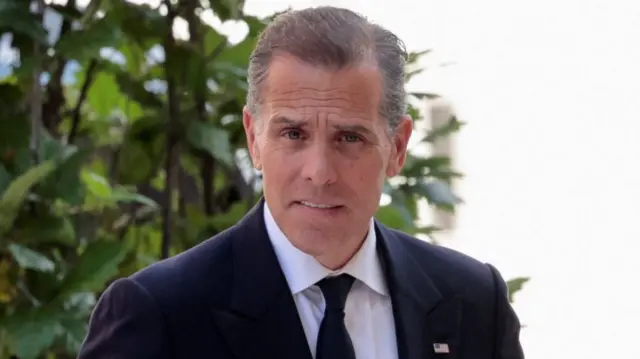 Hunter Biden close-up cutting off shortly below shoulders. He's walking into court wearing a black suit and tie with a white shirt, behind him there's green shrubbery