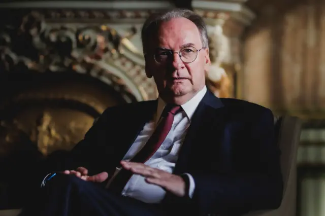 Reiner Haseloff sat in a chair, wearing a suit and red tie with hands raised