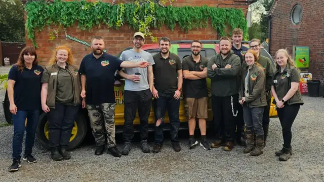 Group shot of 11 staff from Hoo Zoo and Dinosaur World standing in front of a car, all looking at the camera and smiling