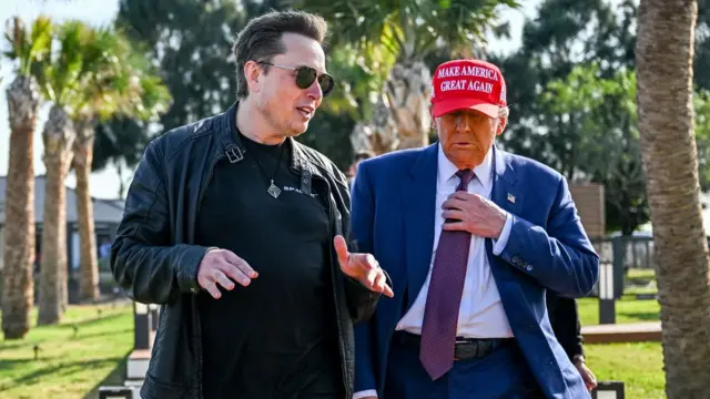 Elon Musk wearing sunglasses talking to Donald Trump who is in a suit and a red Make America Great Again hat, with palm trees in the background