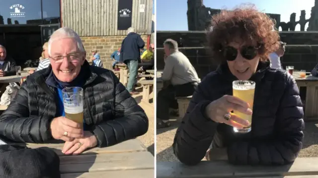 Vince and Josie Byrne drinking pints sat outside a pub. They are both wearing black puffer jackets and holding their drinks while smiling