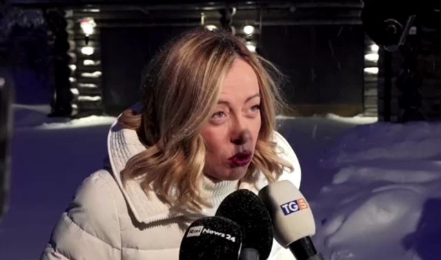 Meloni speaking into several press microphones as she wears a white winter coat and stands in front of snow