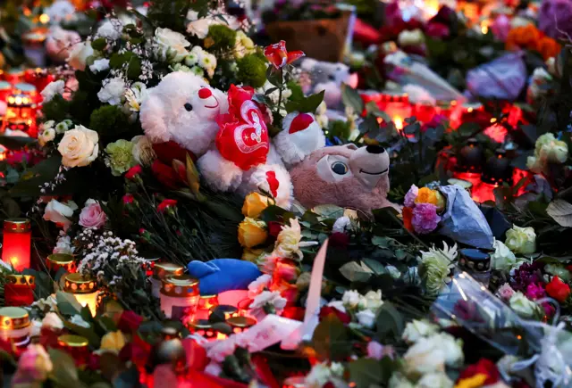 Two teddy bars sit amongst dozens of lit up candles and bunches of flowers