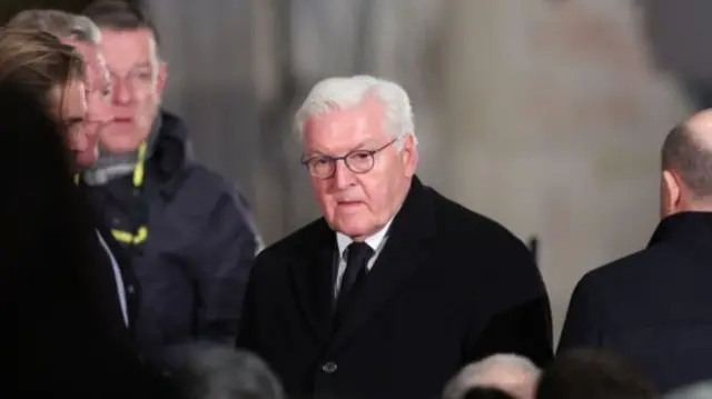 German president Frank-Walter Steinmeier arrives at Magdeburg cathedral for the memorial service. He's wearing a black winter coat and black tie. To his left is Olaf Scholz with his back turned. Members of his entourage and an emergency respondent stand to his right