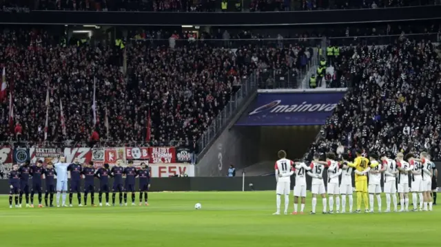 Wide shot showing two football teams opposite each other, each team standing in a line with arms linked and heads bowed with crowded stadium in background