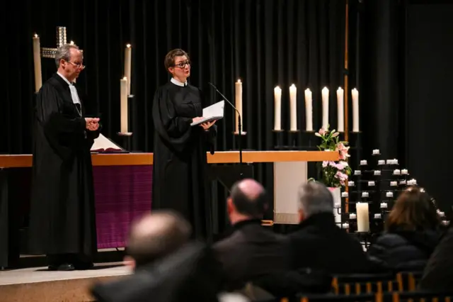 Pastor Sarah-Magdalena Kingreen stands at a microphone in front of a church alter with candles, seated parishioners blurred in front of her