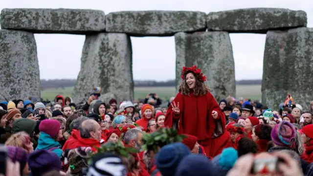 A woman dressed in red and wearing a flower crown sits on someone's shoulders amid a crowd of people at Stonehenge