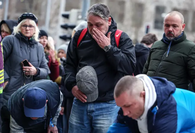 A man wipes a tear from his face as he looks on to flowers being laid