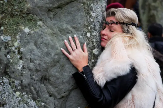 A woman hugs a rock. She wears a furry shawl and a headband