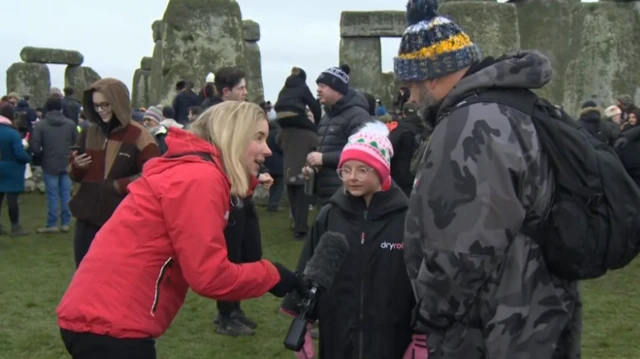 A reporter holds a microphone to a young girls face, who wears a beanie and a dryrobe. A man in stood next to the girl, facing her, also in a dryrobe and beanie