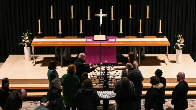 Group of people line up to light candles in front of a raised altar in a church, a large cross sits in the middle of the altar