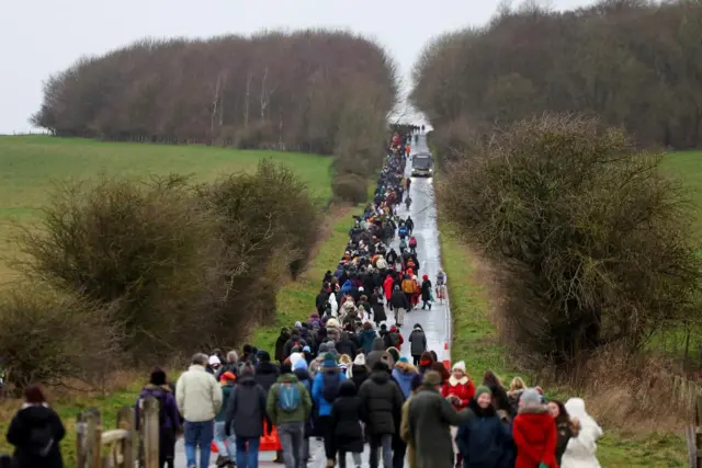 A long crowd of people walks up a road between two hedges and fields