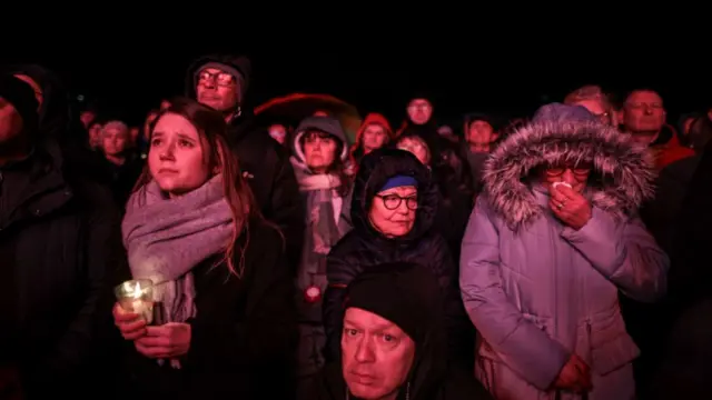 Magdeburg residents attend vigil for the victims of the Christmas market attack. To the left, a girl in a black coat and grey scarf holds a candle, two older women in winter gear to her left. A man in a black hat is visible at the centre-bottom of the frame. A large crowd visible in the background