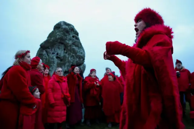 A group of women wearing red coats and clothing gathered round in a semi circle. It appears that they are singing