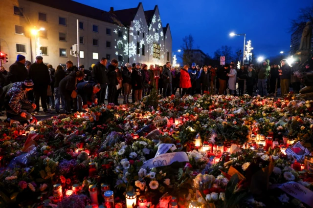 Hundreds of candles and flowers glow in the dark as dozens stand around the tributes