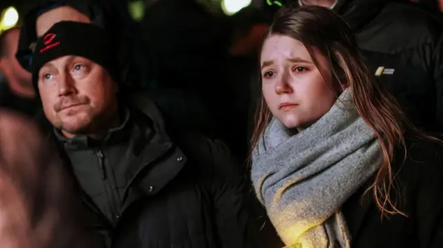 A woman, with large grey scarf, holds a candle with a furrowed brow. A man, with a beanie and winter jacket, stands next to her