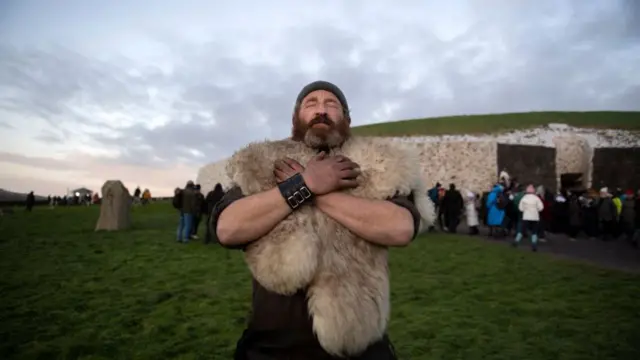 A man wearing a fur-like outfit stands in front of the Newgrange monument - a large mound with a stone facade and grass on top.