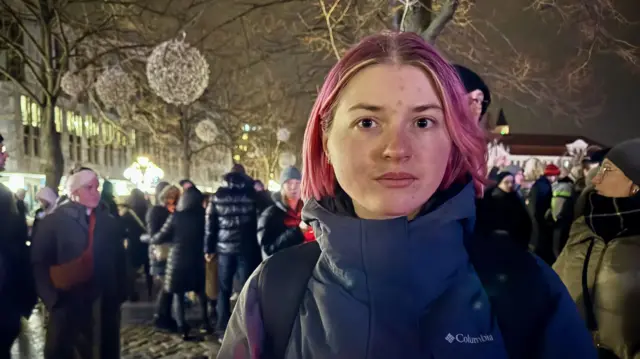 Ukrainian student Tatiana attends memorial service in Magdeburg. She has short pink hair and is wearing a blue winter jacket with a black-strapped backpack on her shoulders. Behind her are other locals walking underneath leafless trees