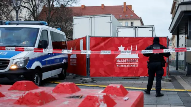 Behind police tape is a police van and an officer standing with their back to a camera in front of a fence for the Christmas market