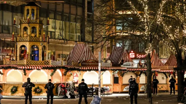 Seven heavily armed police officers in black uniform guard the perimetre of the Magdeburg Christmas Market after a car ploughed into the crowd. Behind the officers are brightly lit stalls and trees covered in Christmas lights