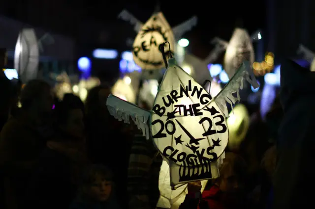 A cream coloured paper lantern with a picture of clock hands, and the words burning the 2023 clocks on it in black