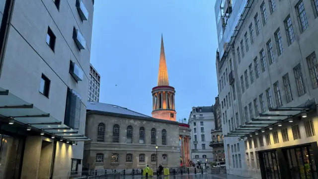 A picture of a plaza between two buildings, and a church in front with the spire lit up. The ground is wet and a security guard stands in the distance