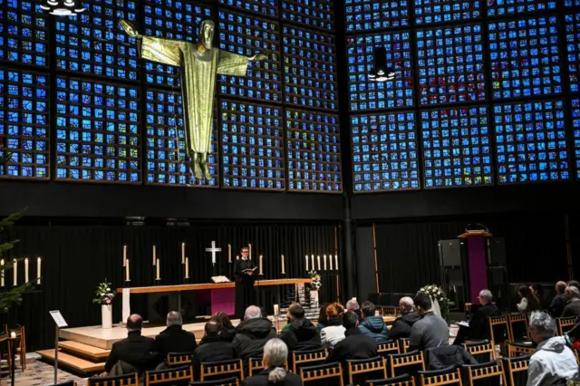 Wide shot of memorial service showing pastor in front of altar, a large man on a cross hanging on the window above the altar and a seated congregation