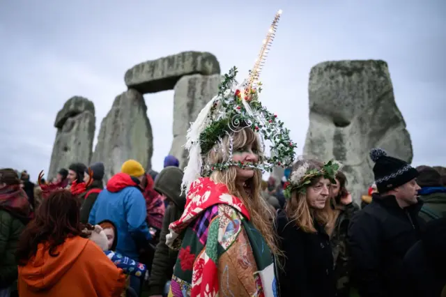 A crowd stood in front of Stone Henge. A woman closest to the camera wears a multicolored cloak and a large flower crown with a horn