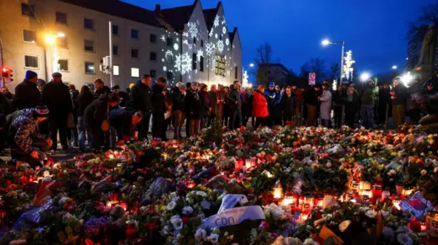 Wide show of flowers and candles on the ground with crowd of people looking, with dark blue night sky and a building with Christmas shaped projections