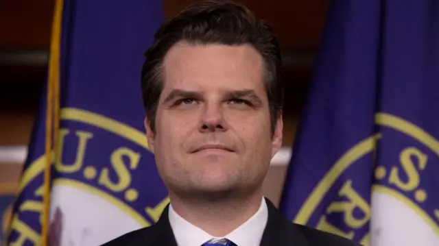 Close-up shot of Matt Gaetz cutting off just below the shoulders. Gaetz is looking straight ahead, behind him are two US Congress flags (blue background with Congress symbol of eagle with US flag draped across it on white background)