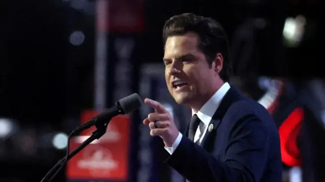 Matt Gaetz speaks at the Republican National Convention in a blue suit behind a podium with a microphone. He points to the left as he speaks.