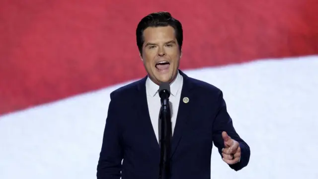 Matt Gaetz stands behind a podium with a microphone in a blue suit, white shirt and tie. He speaks with his hand pointing forward. The background is red and white.