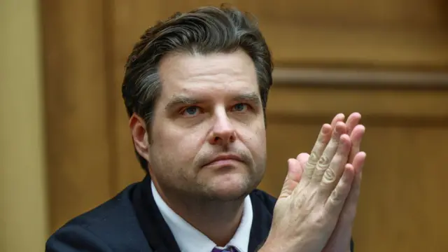 Matt Gaetz sitting down during committee hearing wearing a black suit and white shirt. His hands are palm to palm in front of him. A blurred wood-panelled wall is visible behind him