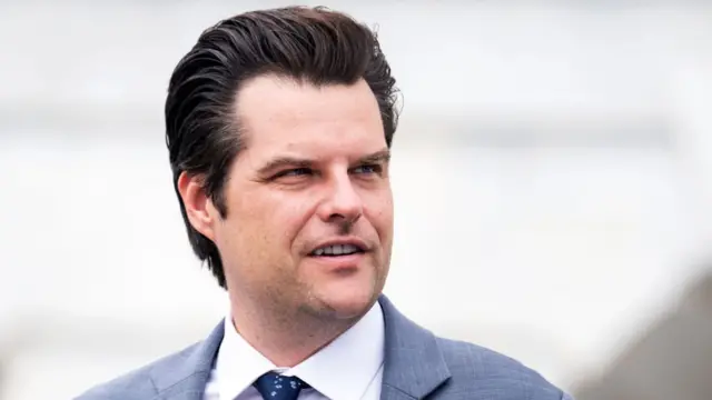 Close up shot of Matt Gaetz cutting off below the shoulders. He's wearing a grey suit with a white shirt and patterned blue tie. The background is a blurry white wall