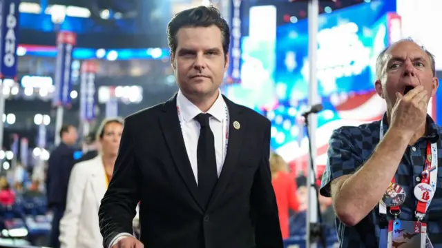 Matt Gaetz in black suit and tie walking around RDC conference. Behind him is the conference hall with attendees and chairs, blurred. A man with a chequered blue and black shirt with his right hand to his mouth stands next to him