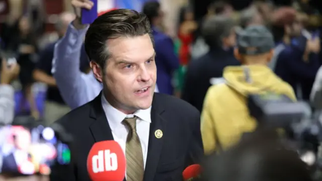 Matt Gaetz answers questions during RNC over the summer. He's in a black suit, gold tie and white shirt as a red mic with DB written on it in white is held towards him. Behind him is a group of people, blurred