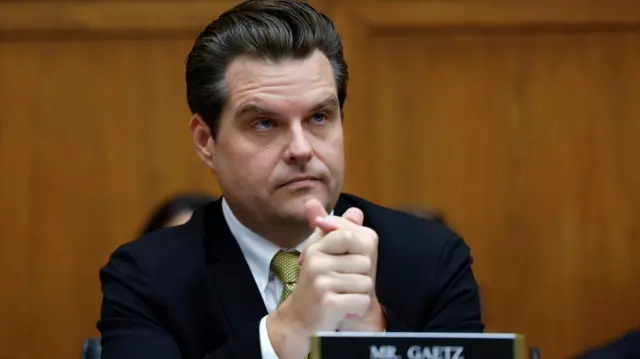 Matt Gaetz sits at a House Judiciary Committee wearing a dark suit with a yellow and black tie. He looks ahead with his hands clasped together.