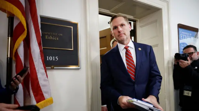 House Ethics Committee chair Michael Guest leaves the committee's main meeting room in Congress. He's in a blue suit, striped red tie and white shirt, and he's holding a notebook and a folder. To the back, to his left, is a man taking a photo of the members leaving