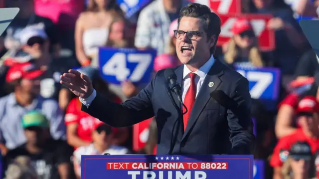 Matt Gaetz speaking at a Trump rally in California during the presidential campaign. He's addressing the crowd from a podium on stage while wearing a suit and red tie. A group of Trump voters is in the background, blurred