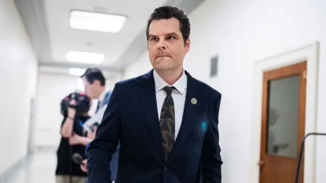 Matt Gaetz walking along a white corridor with bright lights wearing a dark suit, patterned tie and white shirt. A wooden door with a glass panel is visible to his left. Two men are seen standing behind him, one holding a mic and another a camera