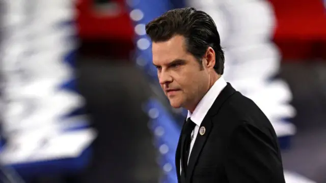 Matt Gaetz mid-shot in black suit, black tie and white shirt at the RNC. Shot is of his left side as he walks, in the background are two rows of chairs blurred