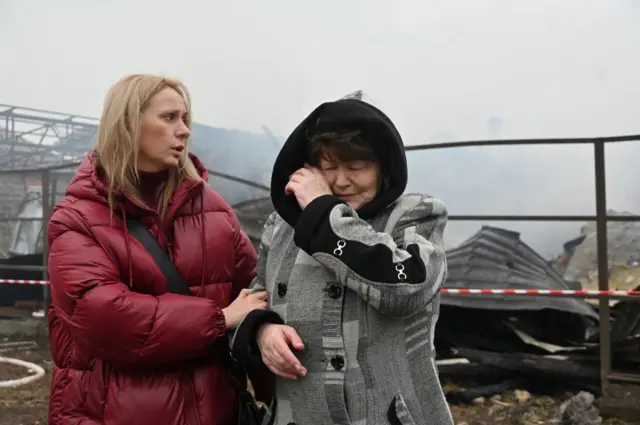 A woman in a grey coat wipes her eye as another - in a red coat - holds her right arm, comforting her. Behind them is damaged homes.