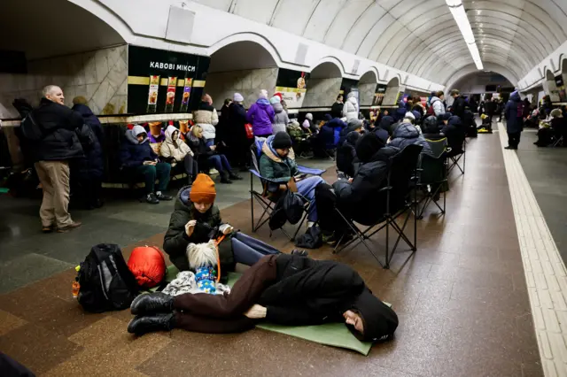 People sitting in chairs and a person in a coat with the hood up, lying down on the floor.