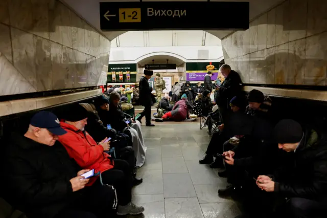 People sitting and standing along the edges of a corridor in a metro station.