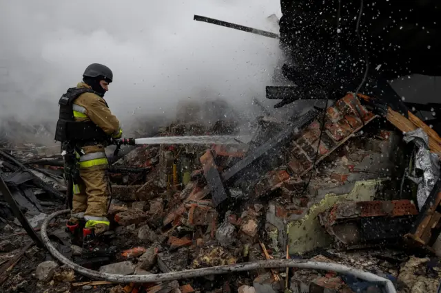 A firefighter using a water hose, aimed at a damaged building blackened by fire and smoke. Smoke is in the air.