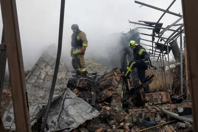 Two firefighters standing in rubble, smoke filling the air