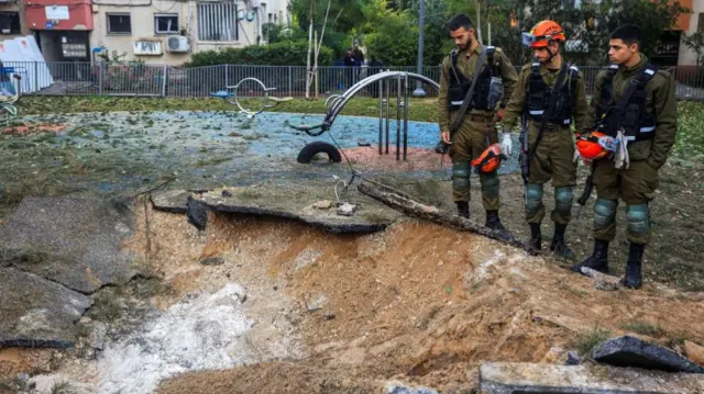 Three emergency services staff in uniform survey a large hole after a strike on a playground in Israel